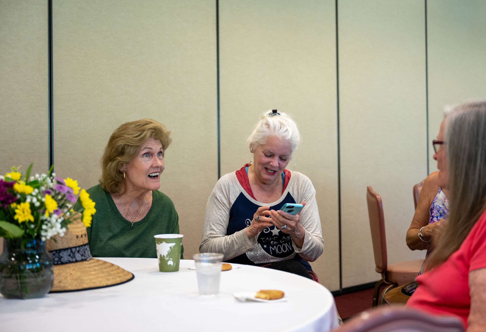 Three older women are sitting at a table laughing and looking at their phones.