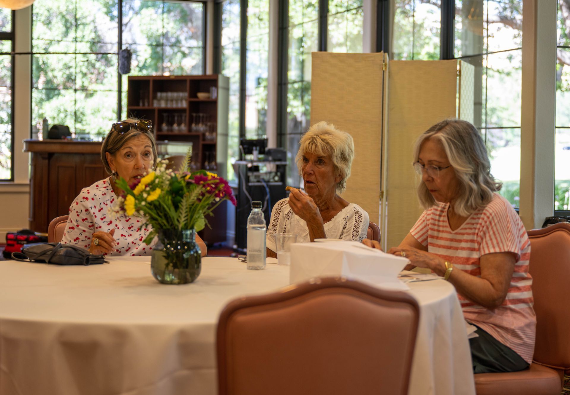 Three women are sitting at a table with a vase of flowers on it.