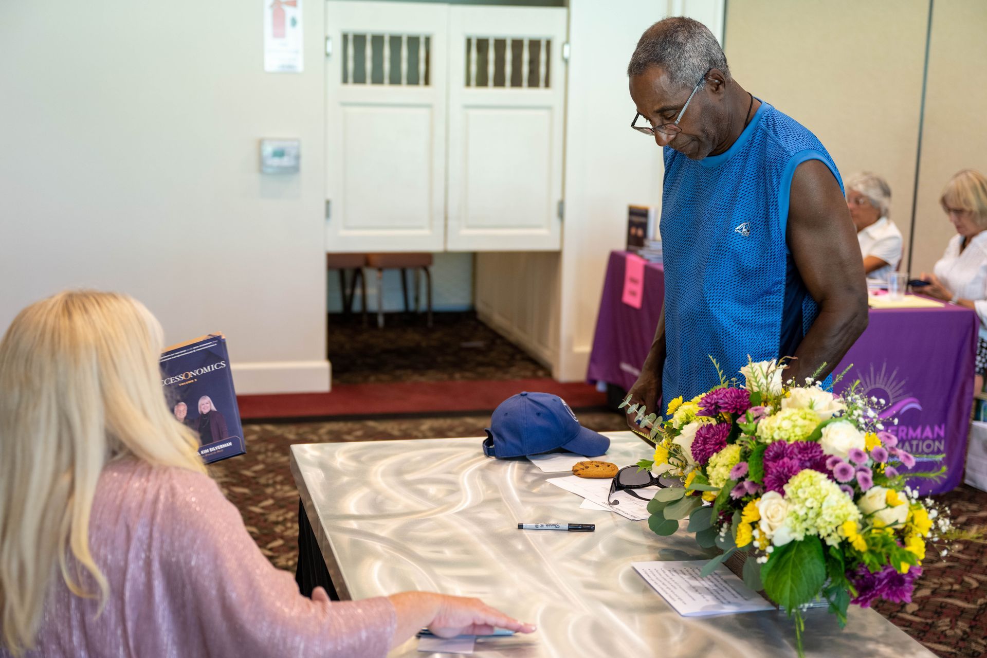 A man is standing and Kim Silverman is sitting at a table with flowers.