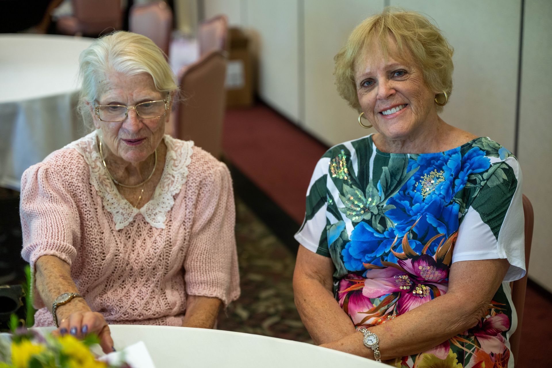 Two older women are sitting at a table smiling for the camera.