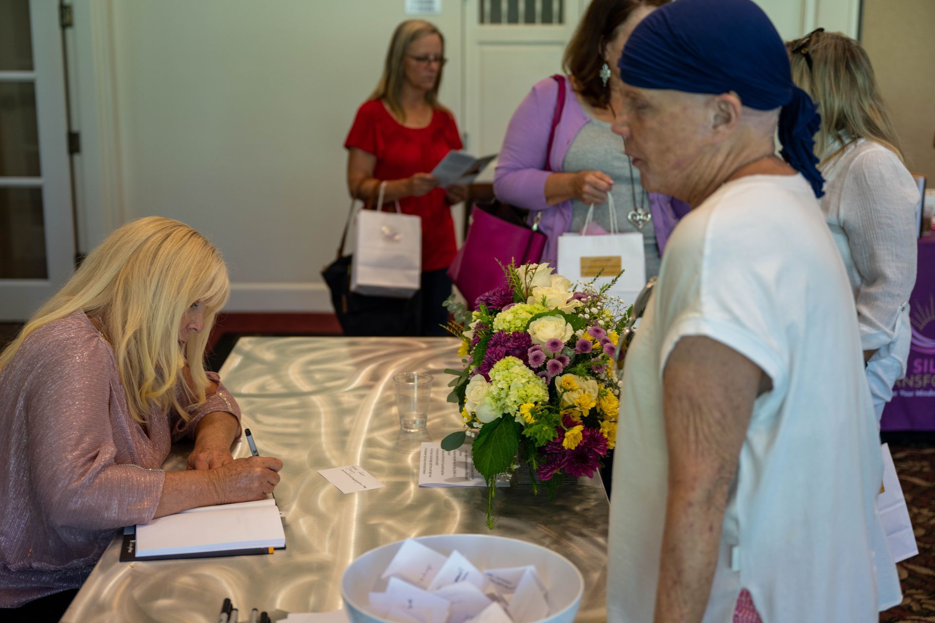 A group of women are standing around a table with flowers waiting for Kim Silverman to sign it.