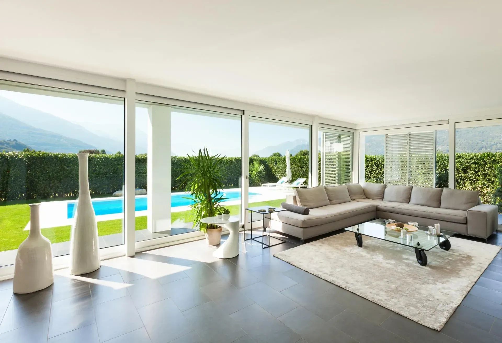 Luminoso soggiorno moderno con finestre a tutta altezza, divano angolare grigio, tappeto bianco e vista sulla piscina.