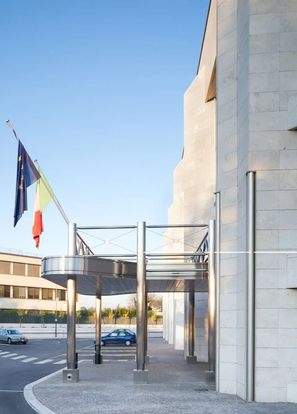 Ingresso di un edificio moderno con tettoia in metallo, bandiere e auto parcheggiate in una piazza soleggiata