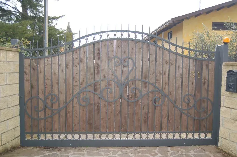 Cancello carrabile in ferro battuto con volute ornamentali davanti a una casa gialla e un muro di pietra.