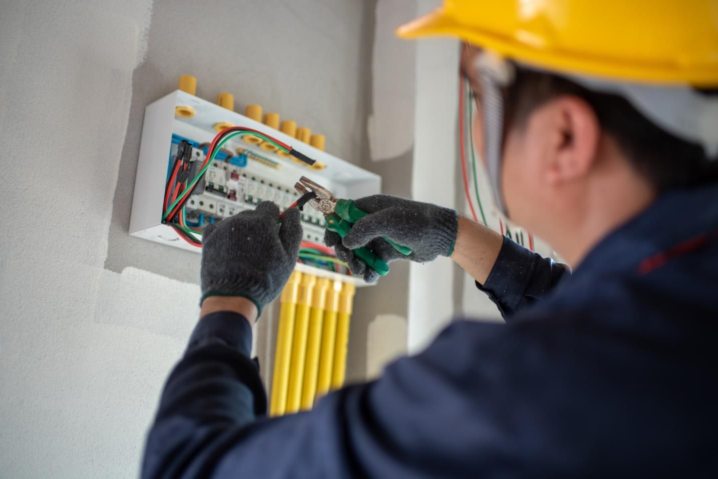 Electrician working on a circuit breaker panel, wearing gloves and a yellow hard hat.