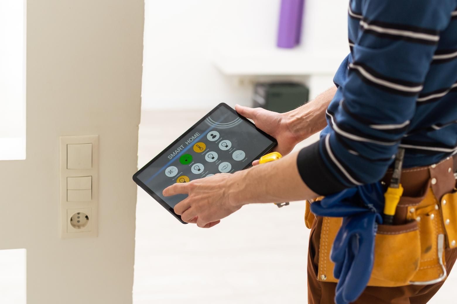 Person using a tablet to control smart home features near a light switch and outlet; tool belt visible.