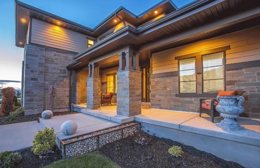 A modern home exterior with stone and wood siding, lit by warm lights. Porch with decorative elements.