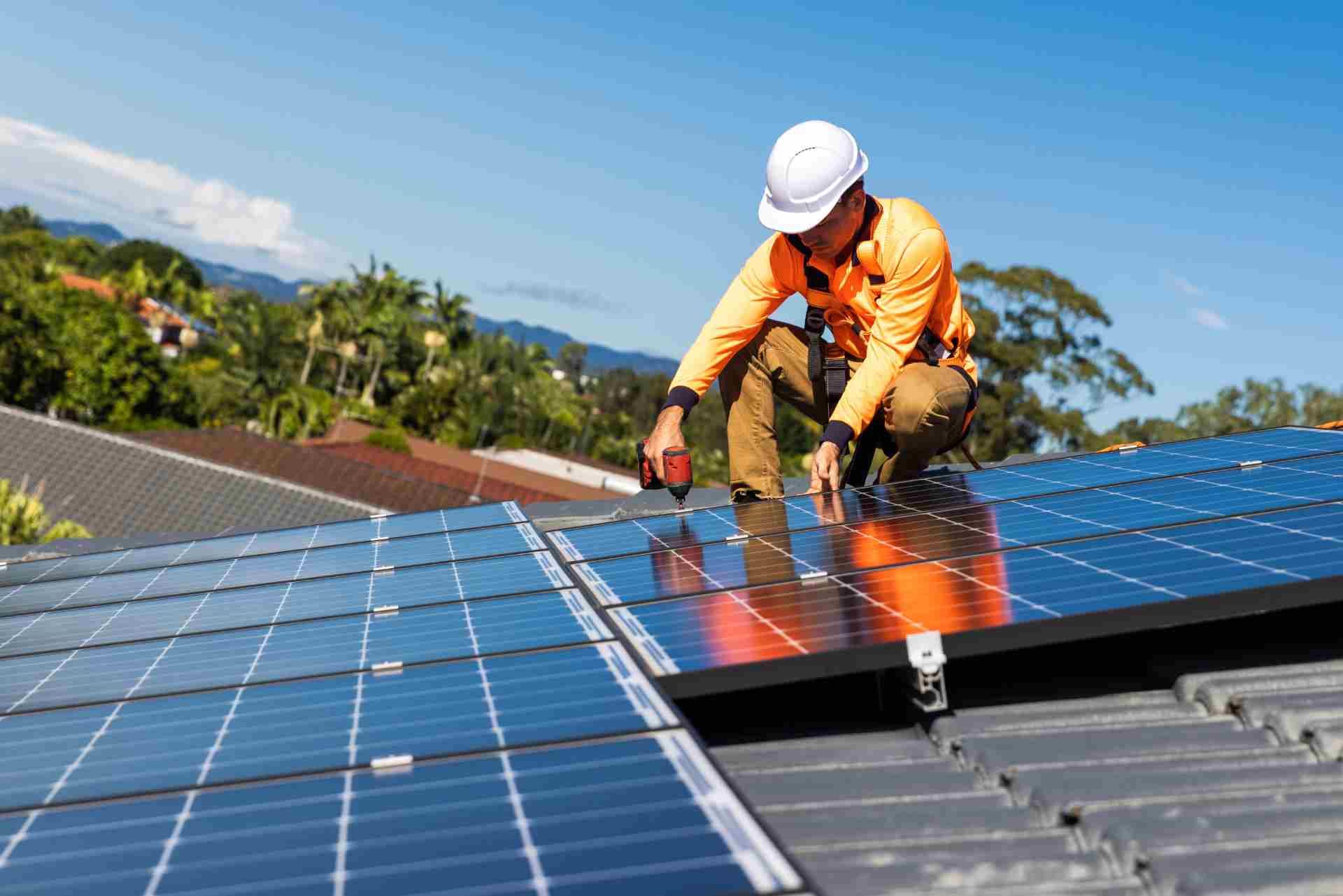 Solar panel installer on rooftop, installing panels with a drill. Bright orange clothing, blue sky.