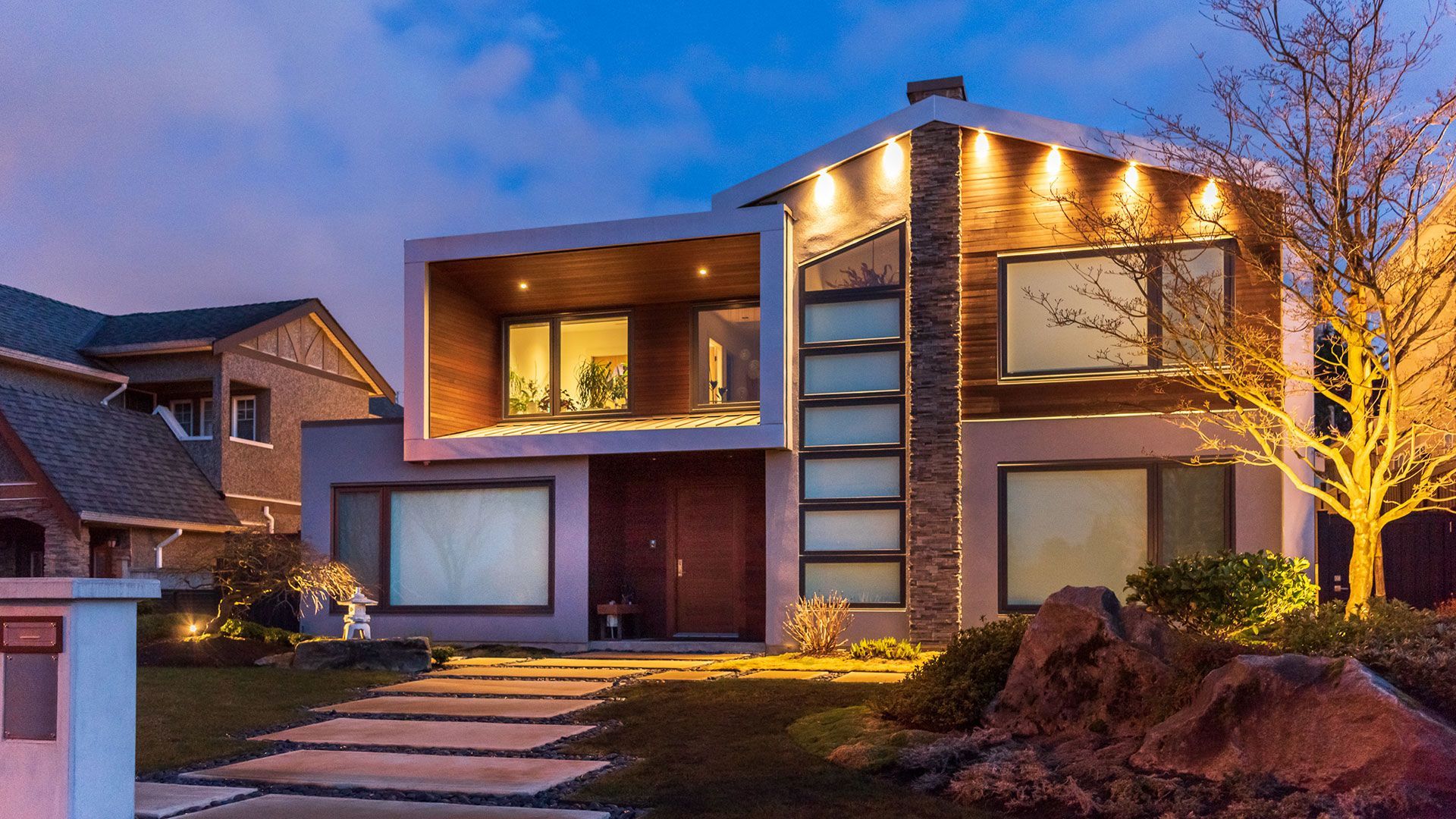 Modern house with square window feature, wooden accents, and a lit pathway at dusk.