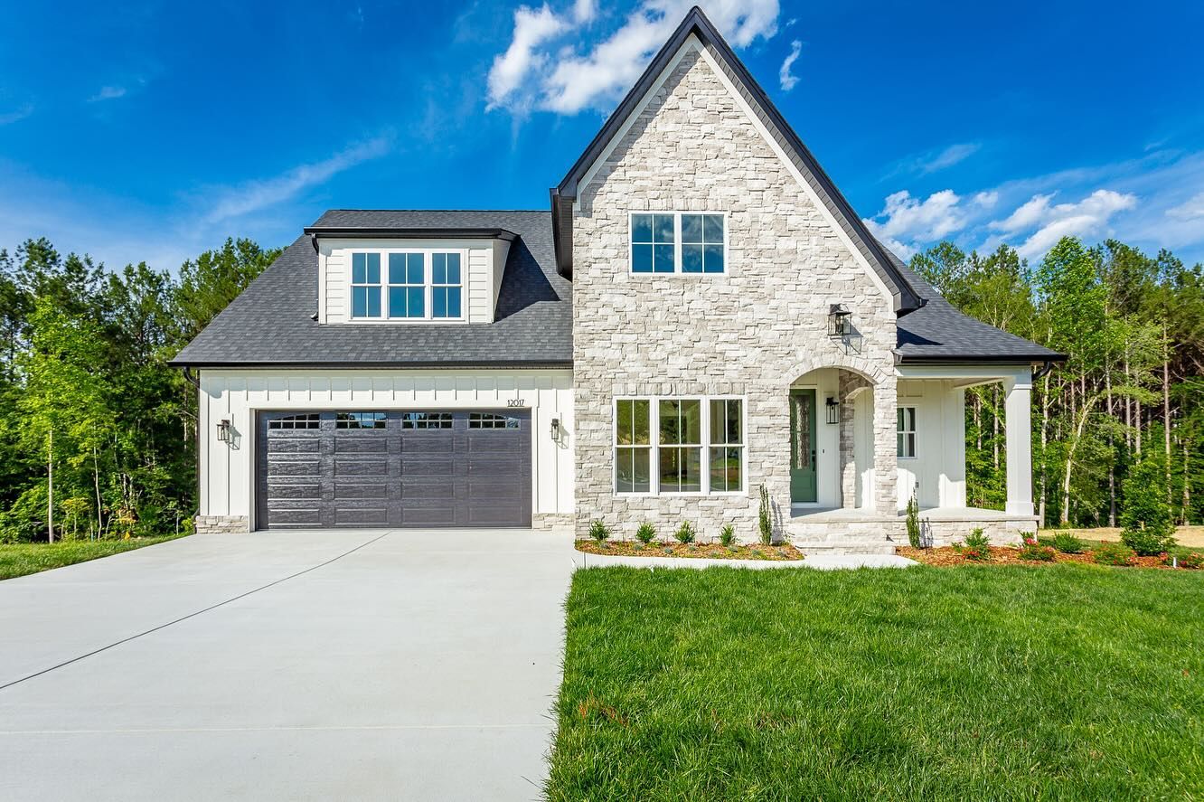 Two-story house with light-colored brick facade, dark garage door, and lush green lawn against a blue sky.