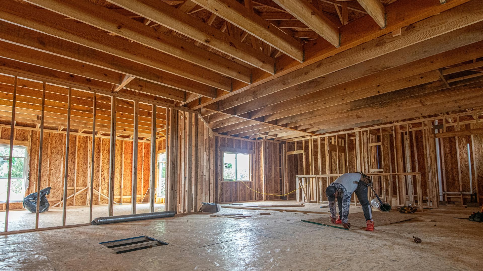 Interior framing of a house under construction, with exposed wooden beams and two workers on a concrete floor
