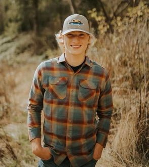 A young man wearing a plaid shirt and a hat is standing in a field.