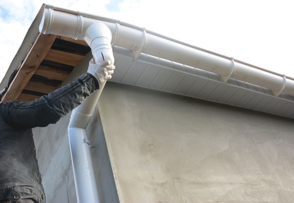 Person Installing a White Gutter and Downspout on a House Roofline — The Roofer Troopers in Wodonga, VIC