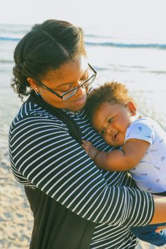 A woman wearing glasses is holding a baby on the beach.