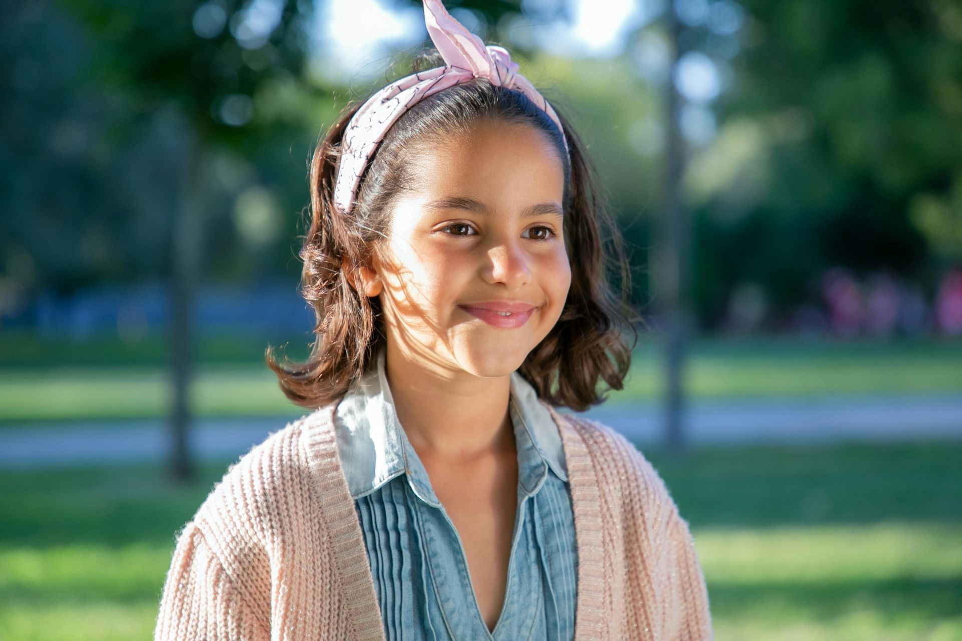 A young girl wearing a headband and a sweater is smiling in a park.