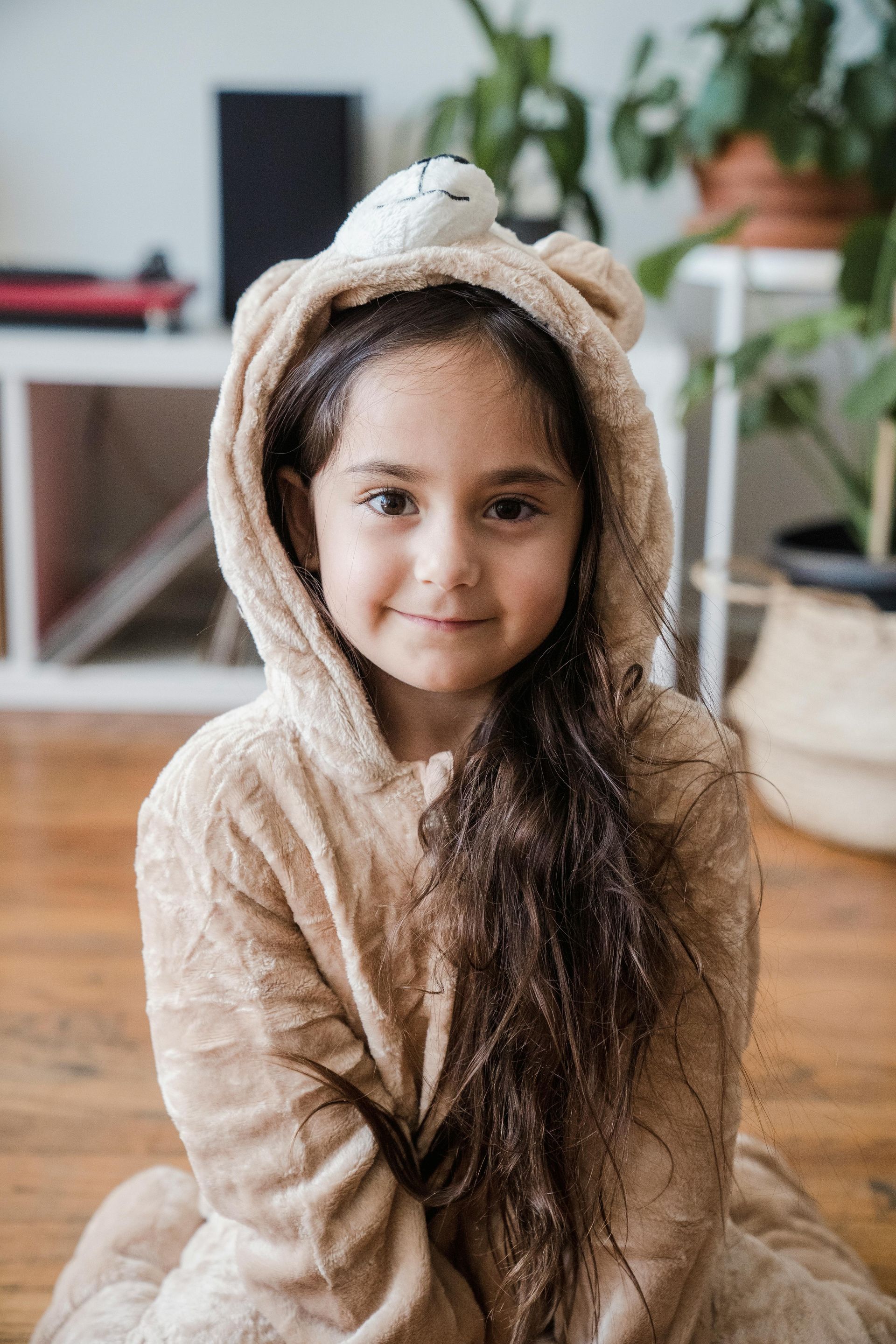 A little girl wearing a teddy bear costume is sitting on the floor.