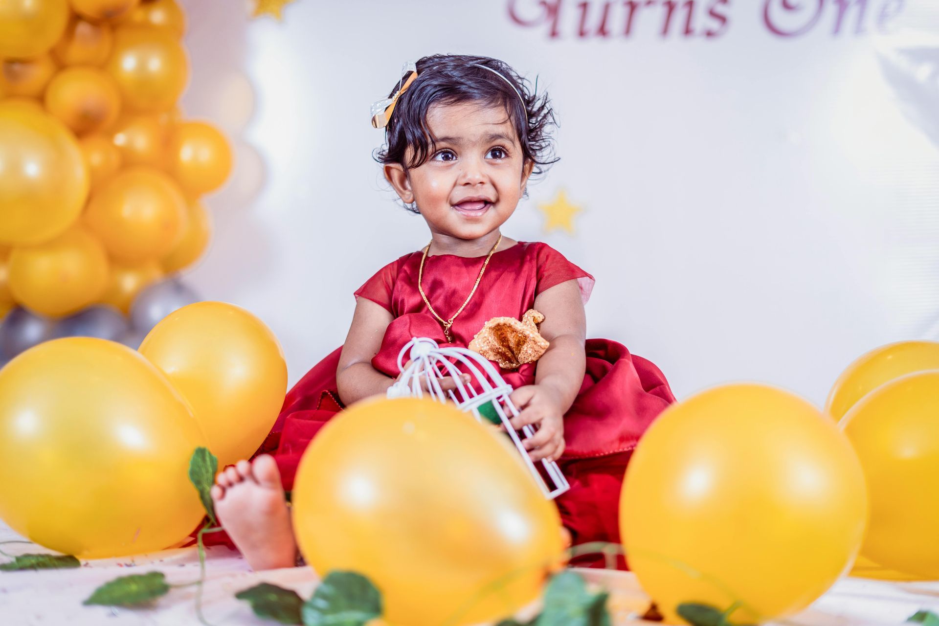 A little girl is sitting on the floor surrounded by yellow balloons.