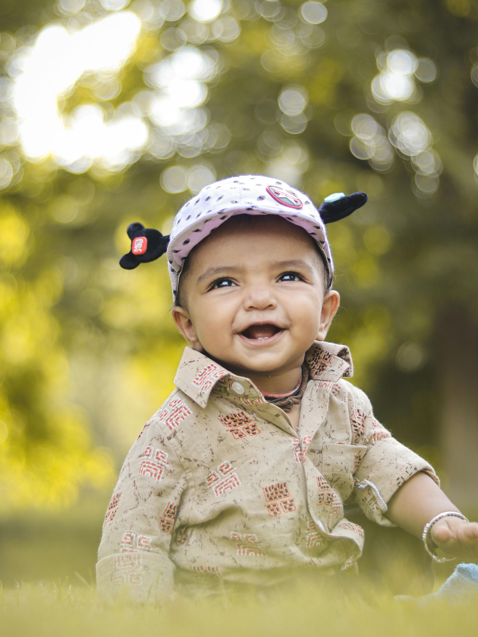 A baby is wearing a hat and smiling while sitting in the grass.