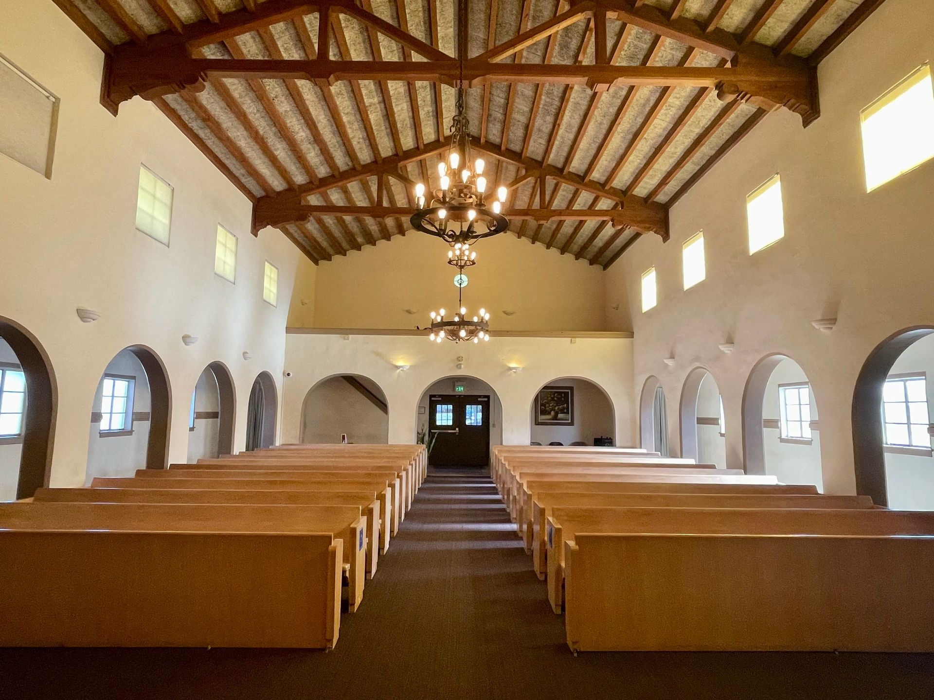 Interior of a chapel with rows of wooden pews, arched windows, and two chandeliers hanging from a wooden beam ceiling.