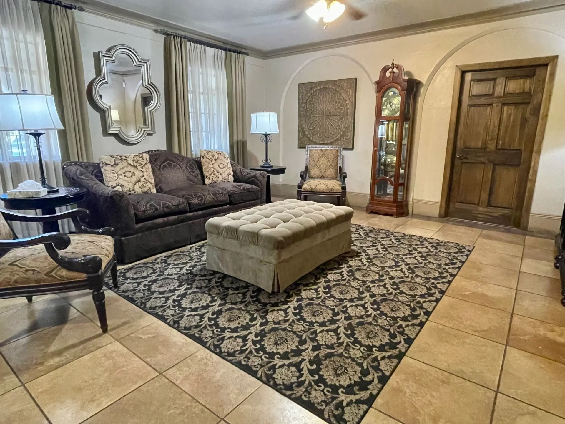 Living room with sofa, rug, ottoman, chairs, mirror, grandfather clock, and beige tile floor.