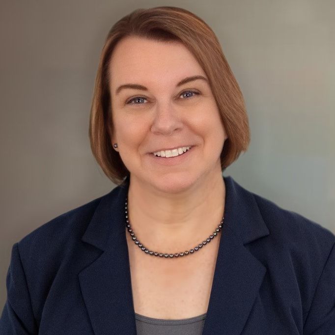 A woman wearing a blue jacket and a necklace is smiling for the camera.