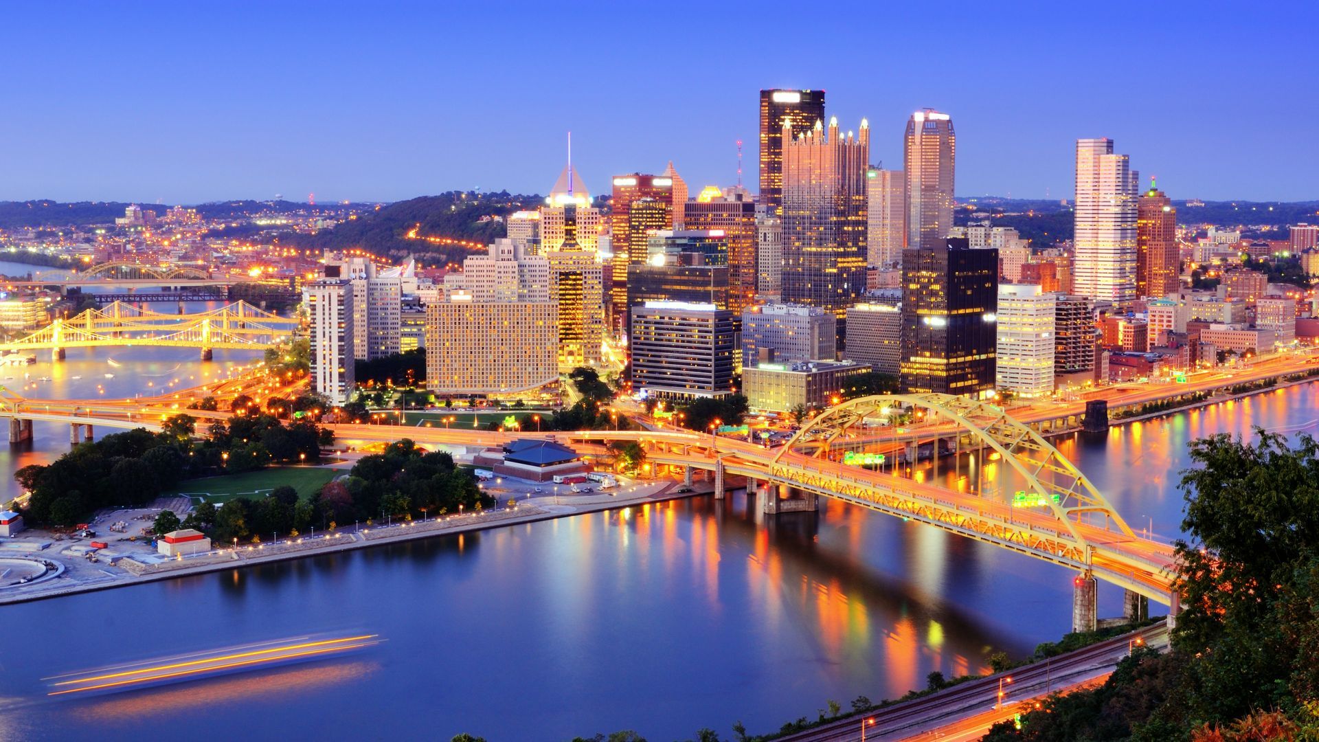 Pittsburgh skyline at dusk, illuminated bridges, river, and buildings with lights.