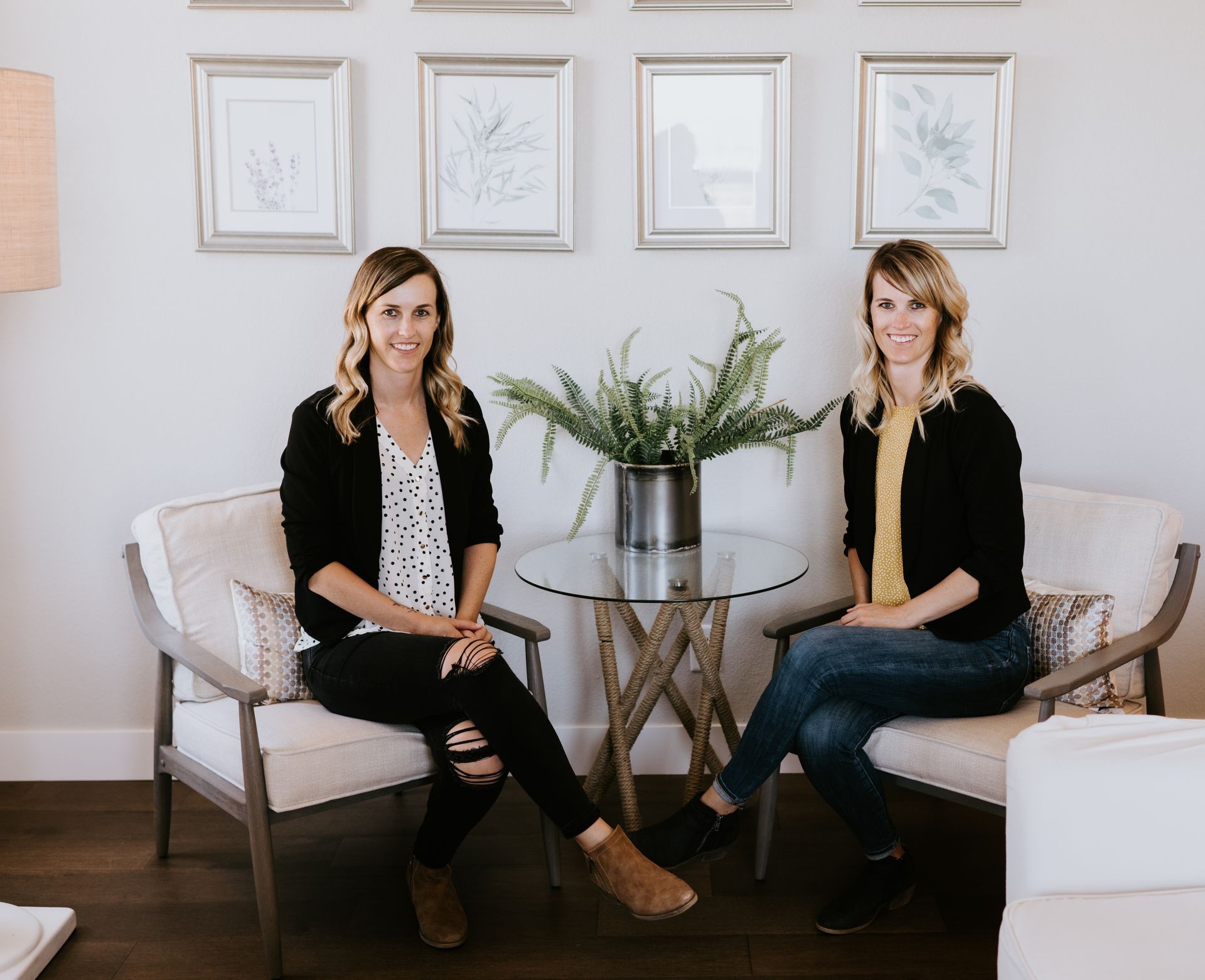 Two people sit in chairs on either side of a small table with a plant, in front of a wall with four framed prints.