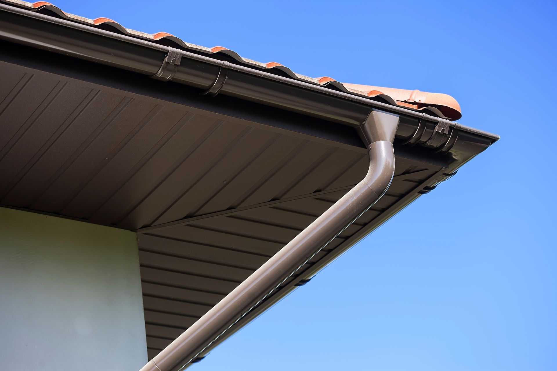 A close-up of a brown roof gutter and downspout installed under a tiled roof overhang against a clear blue sky.