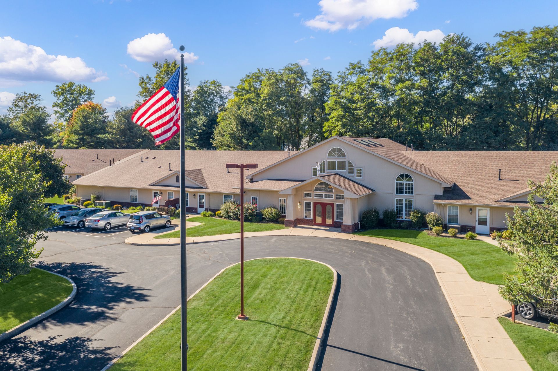 Long, one-story beige building with an American flag, trees, and cars parked in front.