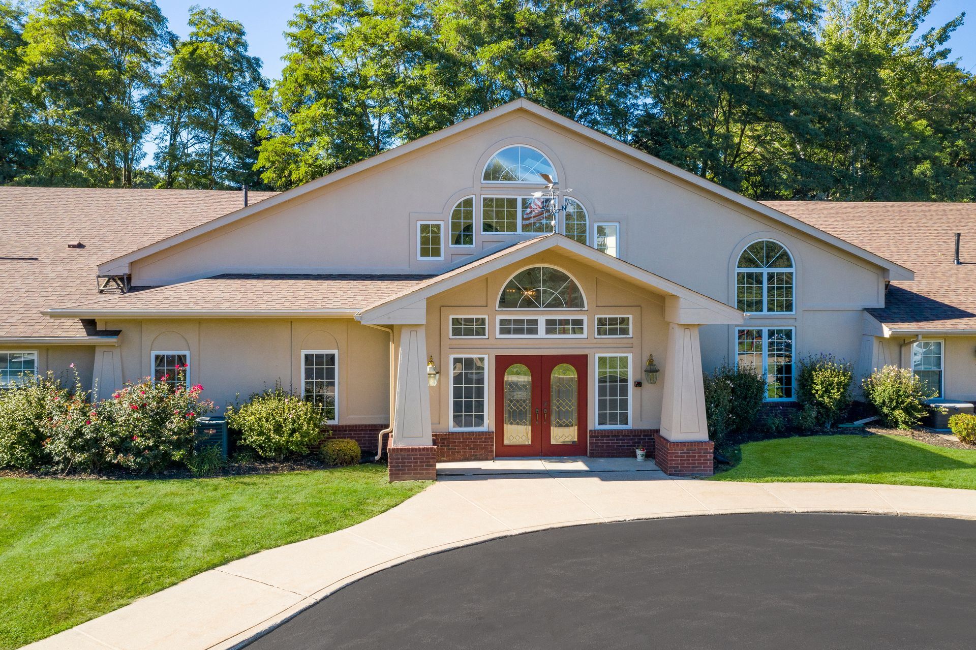 Exterior of a tan building with arched windows, red double doors, and a curved driveway, surrounded by green grass and trees.