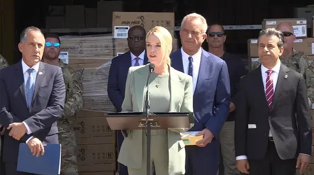 Mujer hablando desde un podio, rodeada de personas con traje y con cajas al fondo.