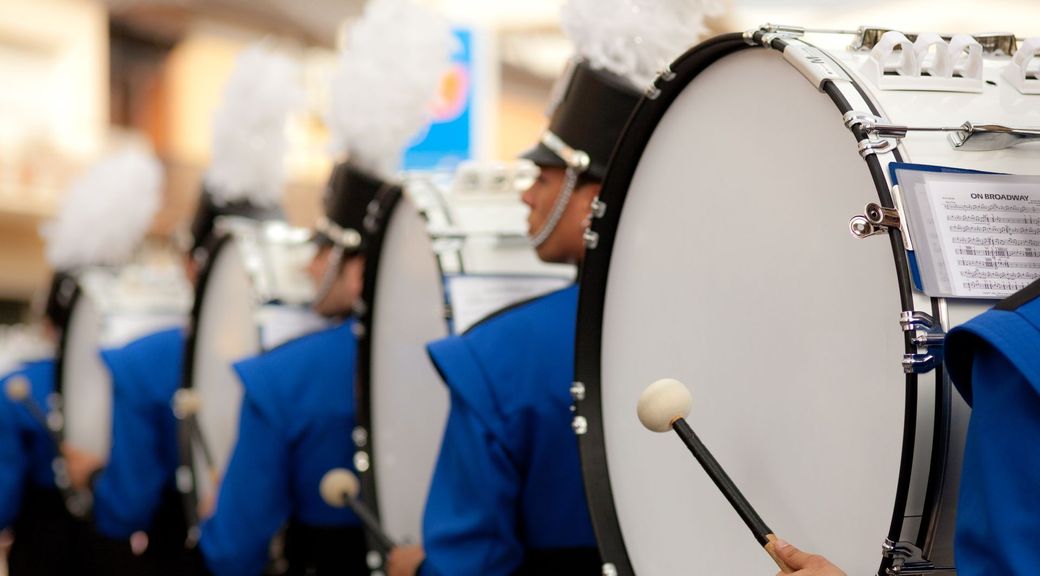 A drummer in a marching band is playing a drum with a mallet.