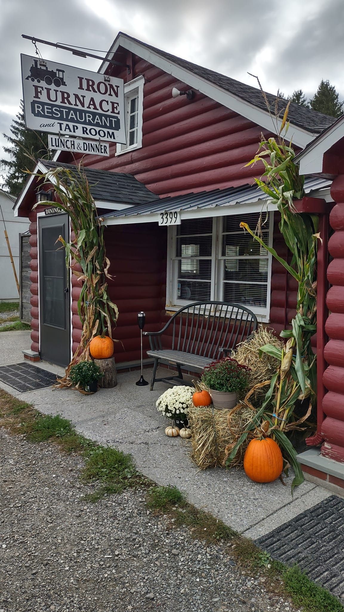 A red log cabin with pumpkins and corn on the cob in front of it.