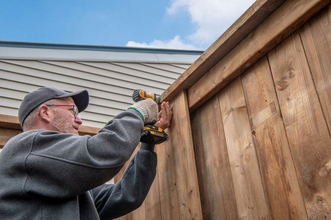 Person using a drill to build a wooden fence outdoors under a blue sky