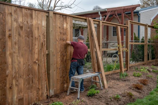 Person installing a wooden fence in a backyard garden, standing on a step ladder.