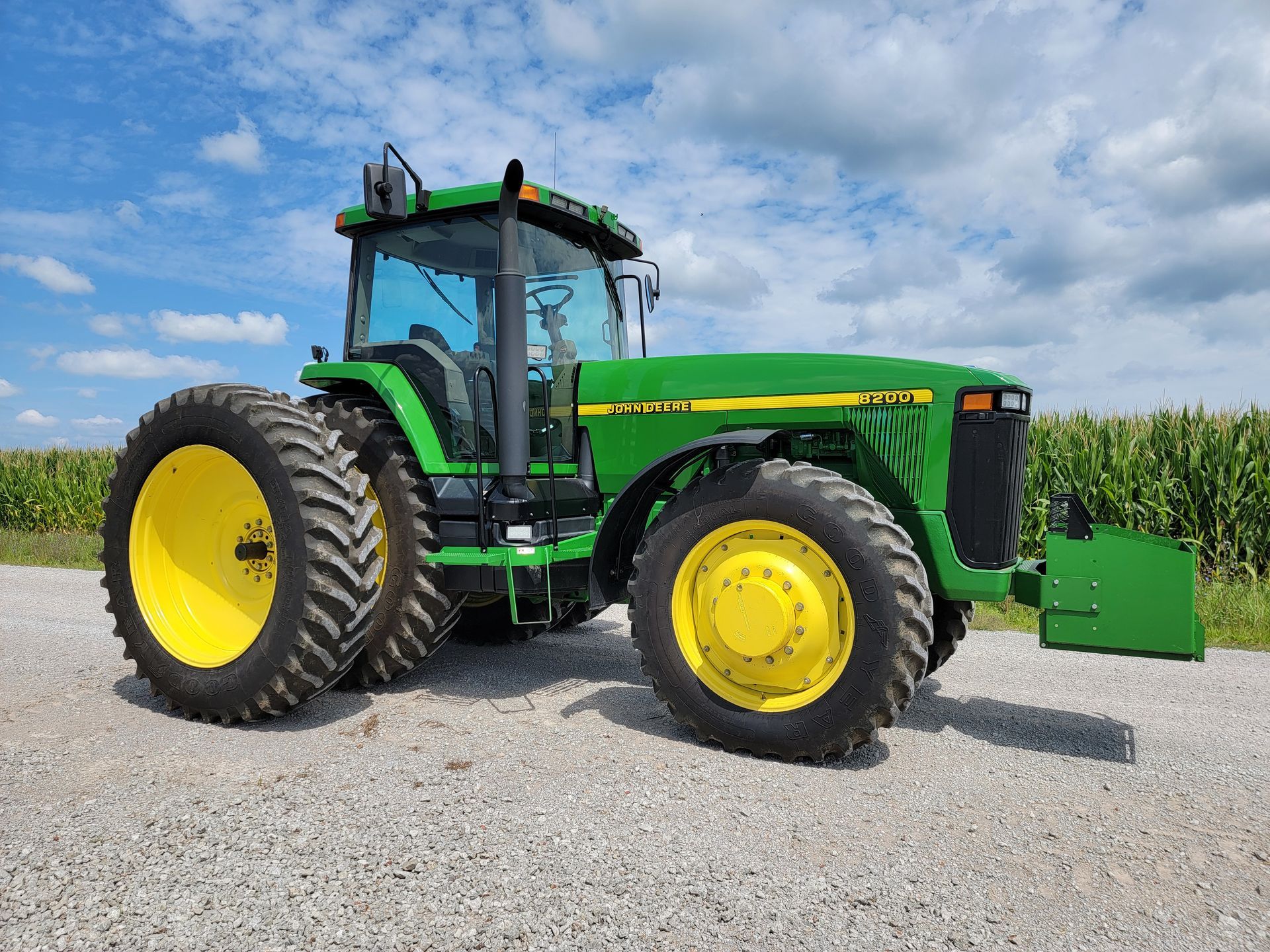 Green and yellow John Deere tractor on a gravel path near a cornfield under a blue sky.