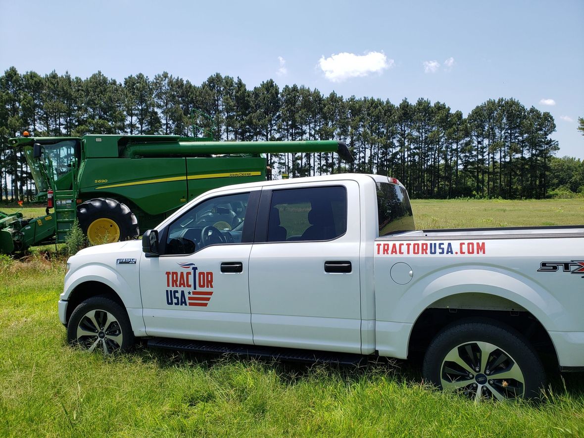 White Ford truck with Tractor USA logo parked in a field, John Deere combine in the background.