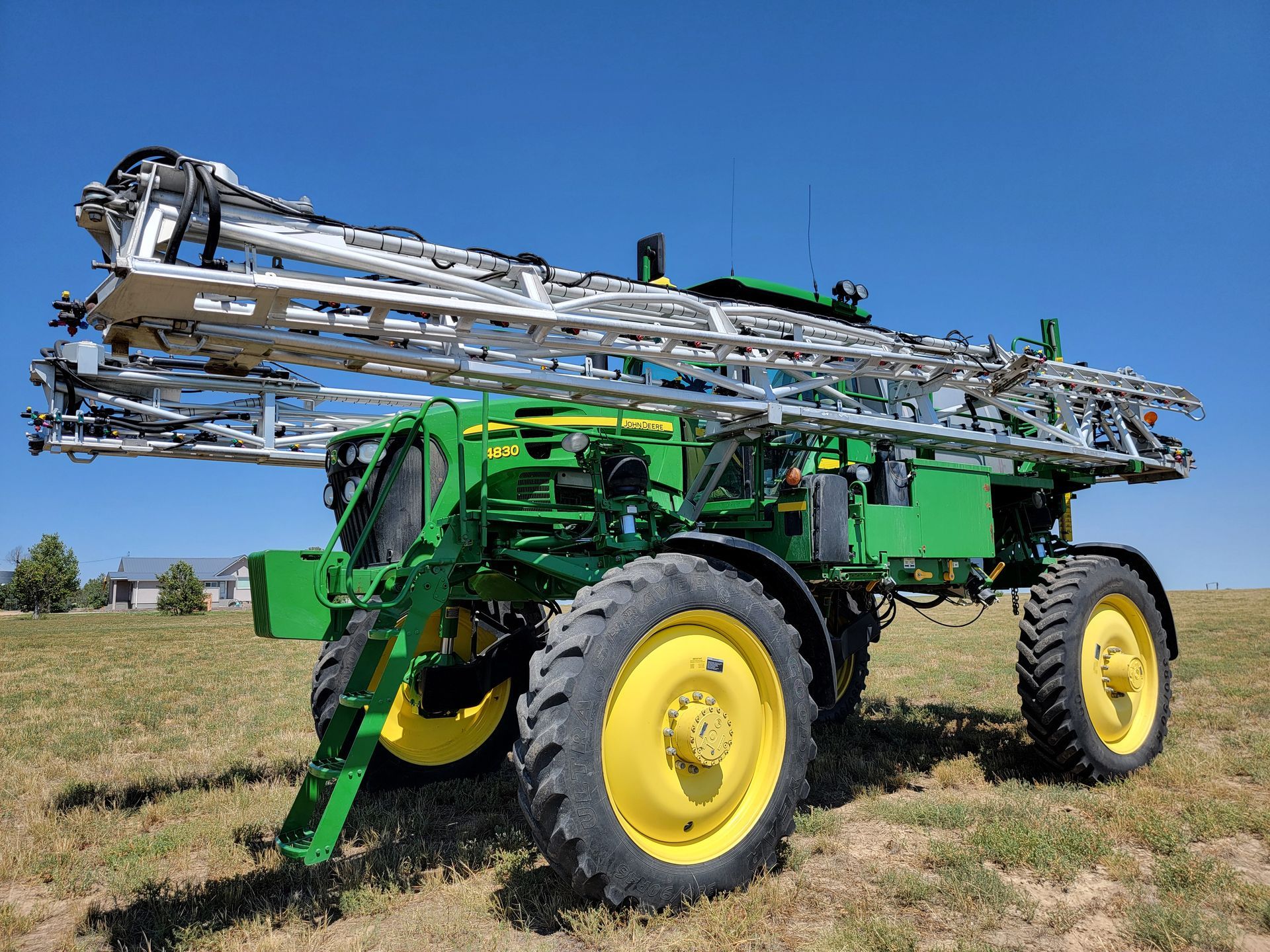 John Deere agricultural sprayer on a grassy field, under a blue sky. Green and yellow tractor with extended booms.