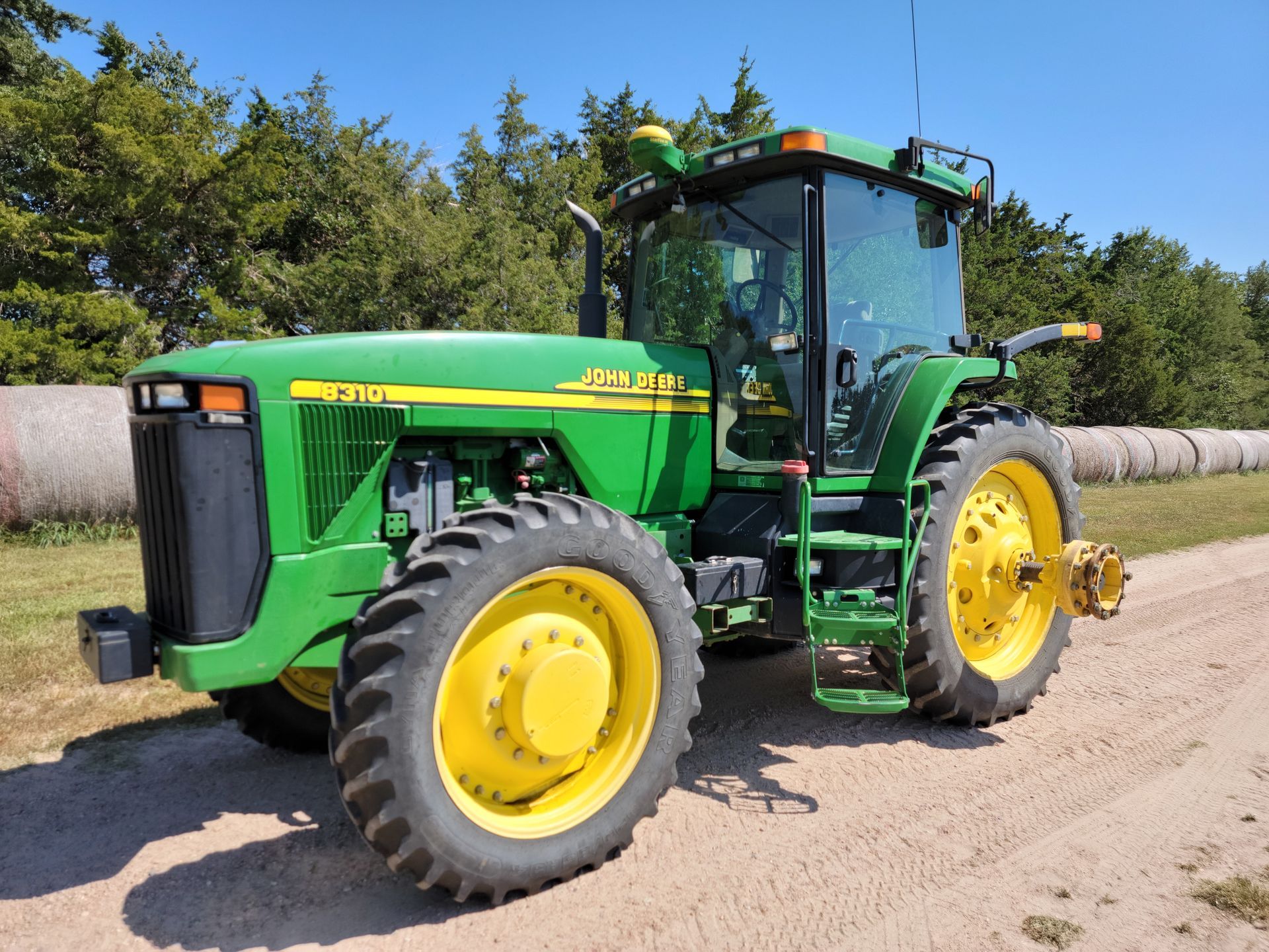 Green and yellow John Deere tractor on a dirt road, next to hay bales and trees.