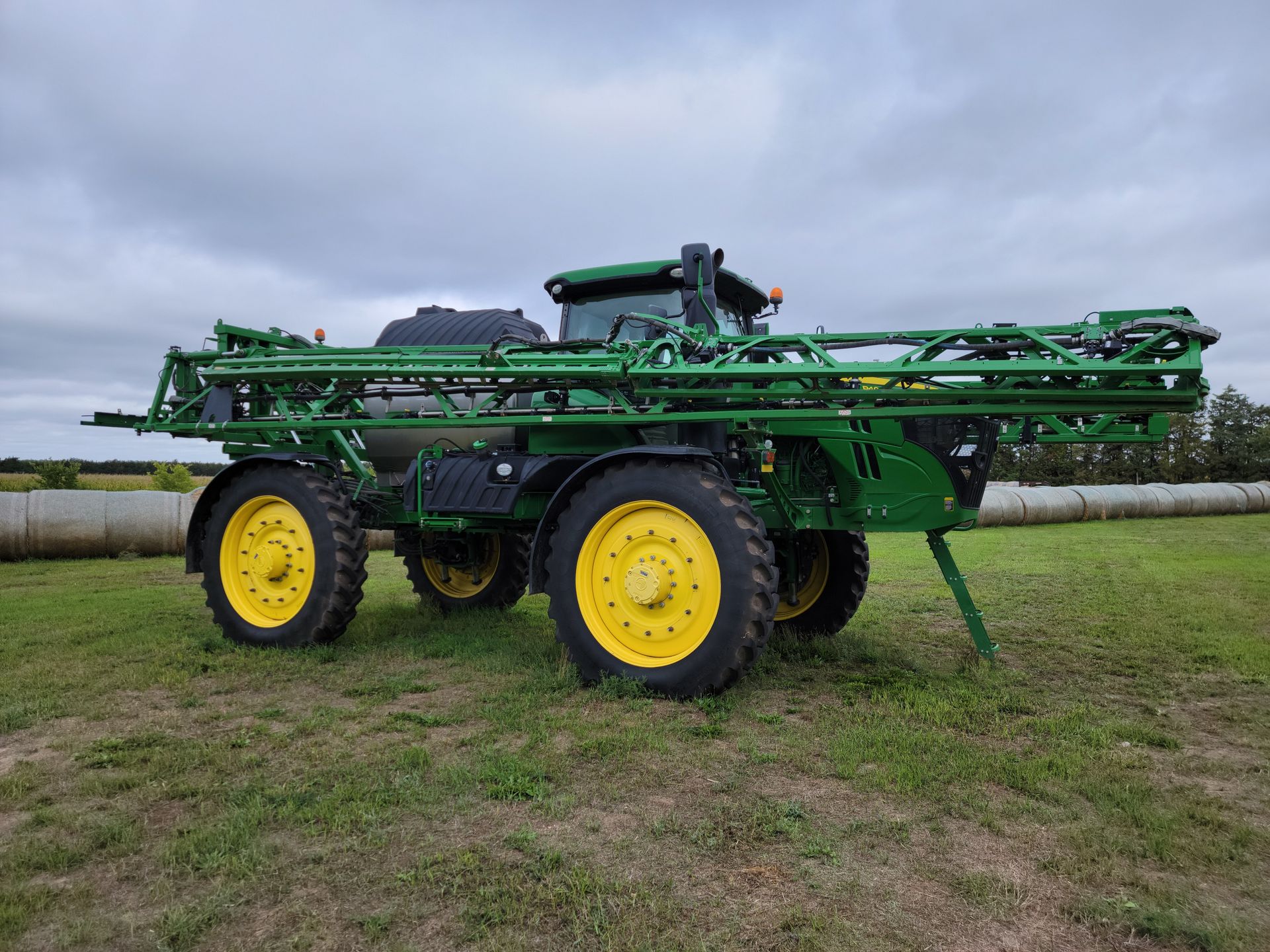 Green and yellow John Deere agricultural sprayer on a grassy field under a cloudy sky.