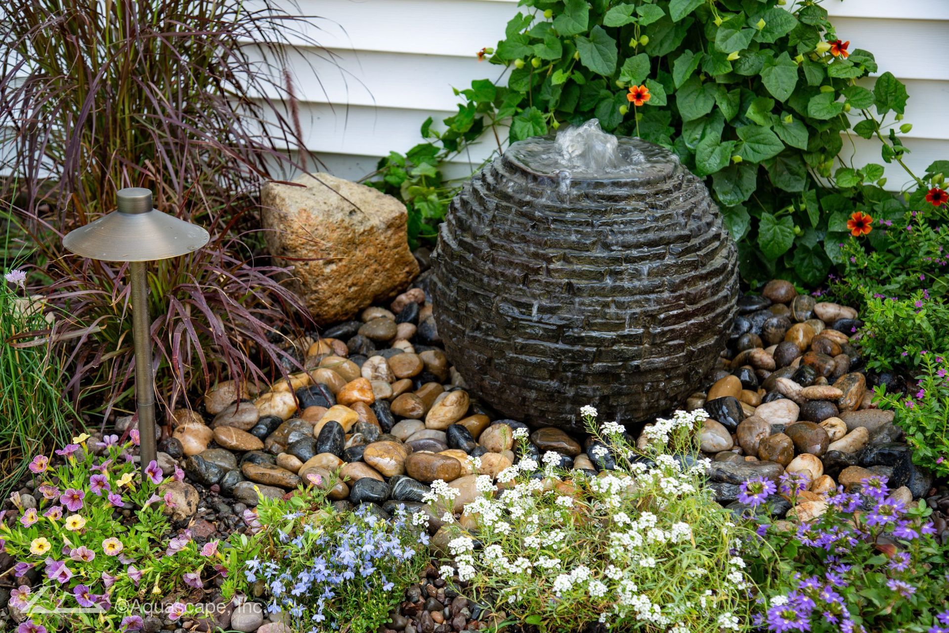 A water fountain surrounded by colorful flowers and river stones in a garden setting.