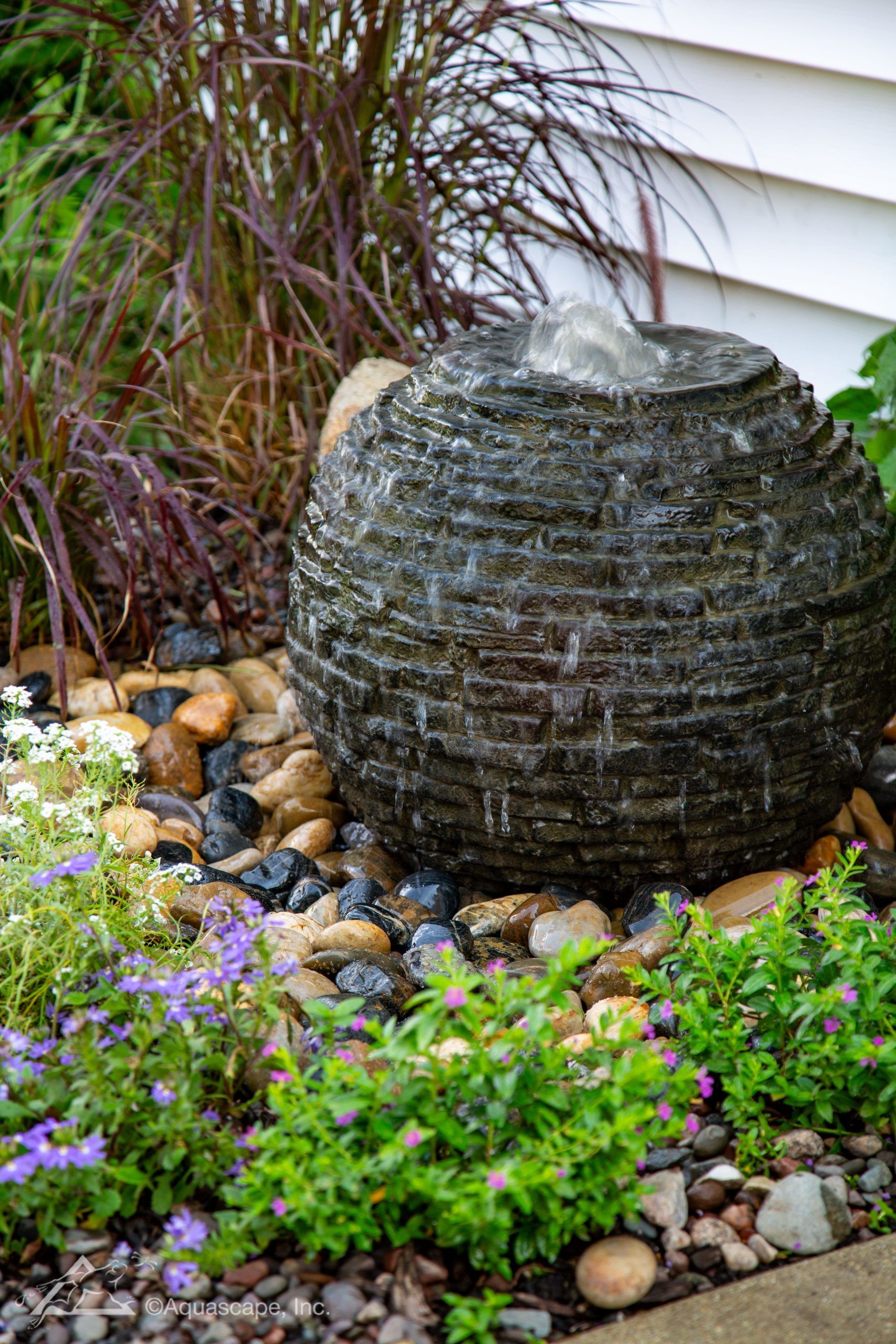 Spherical water fountain in garden with rocks, purple flowers, and red ornamental grass.