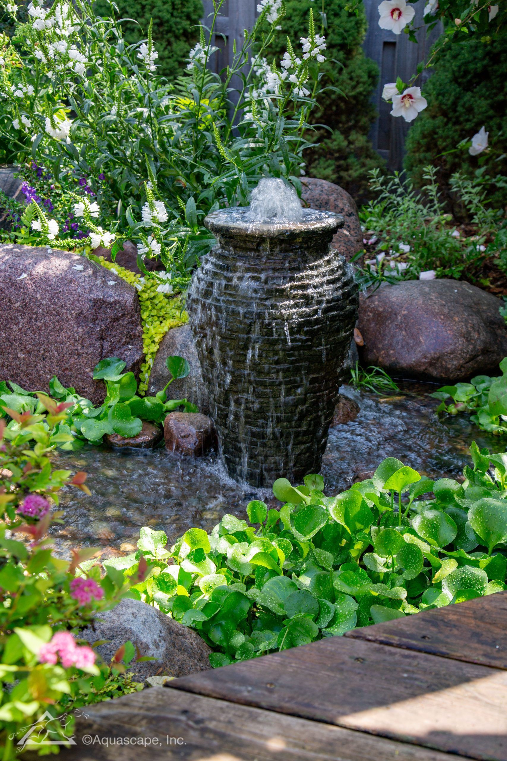 Water fountain cascading into a pond with lush greenery and large rocks.