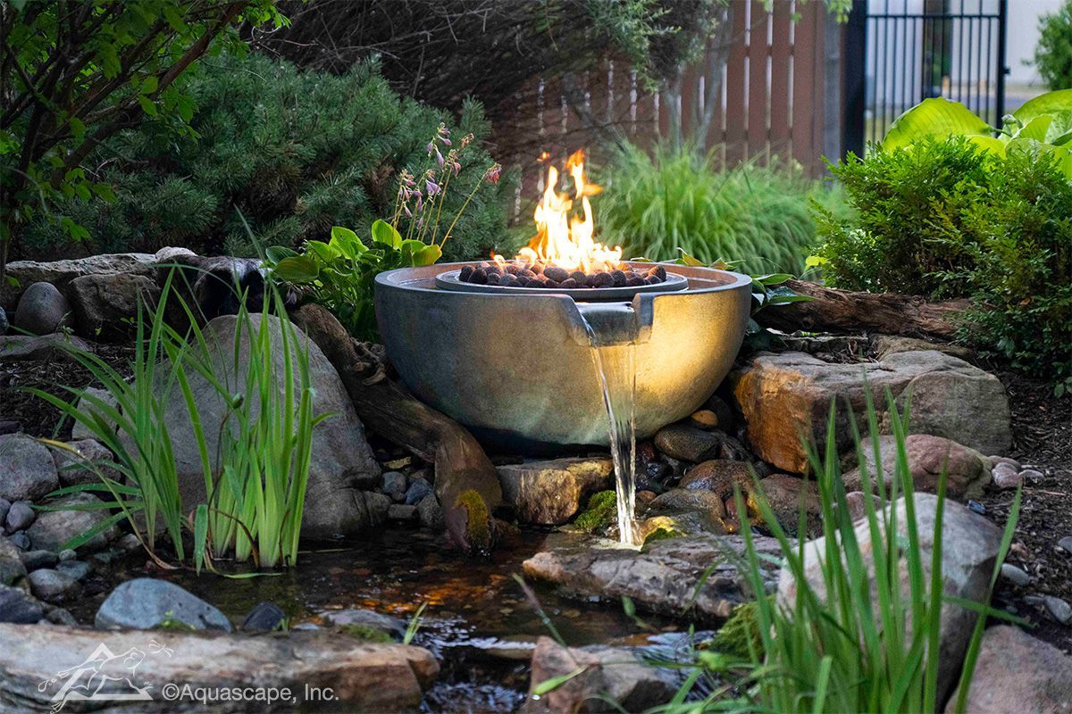 Fire pit with flowing water feature in a garden, surrounded by rocks and greenery.