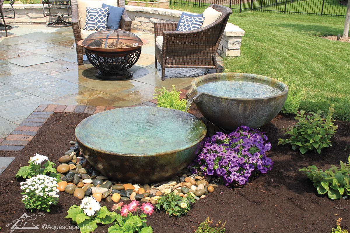 Outdoor water fountain with turquoise bowls, surrounded by flowers and rocks. Patio with seating in background.