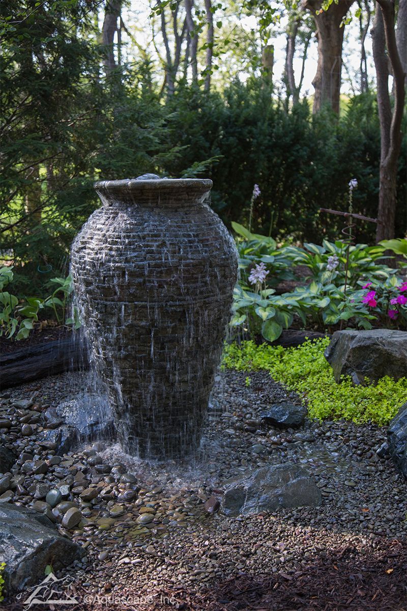 Stone urn water fountain in a garden setting, water cascading down, surrounded by greenery and plants.