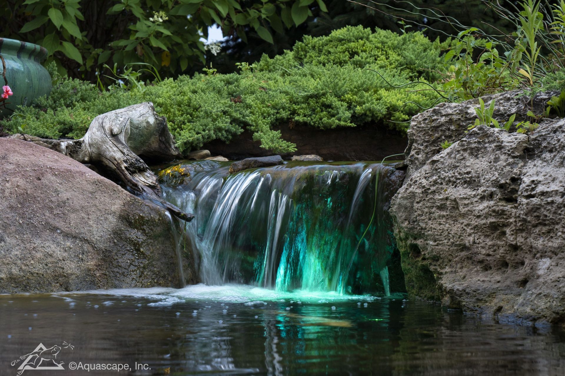 A small waterfall in a pond, with green illuminated water, surrounded by rocks and greenery.