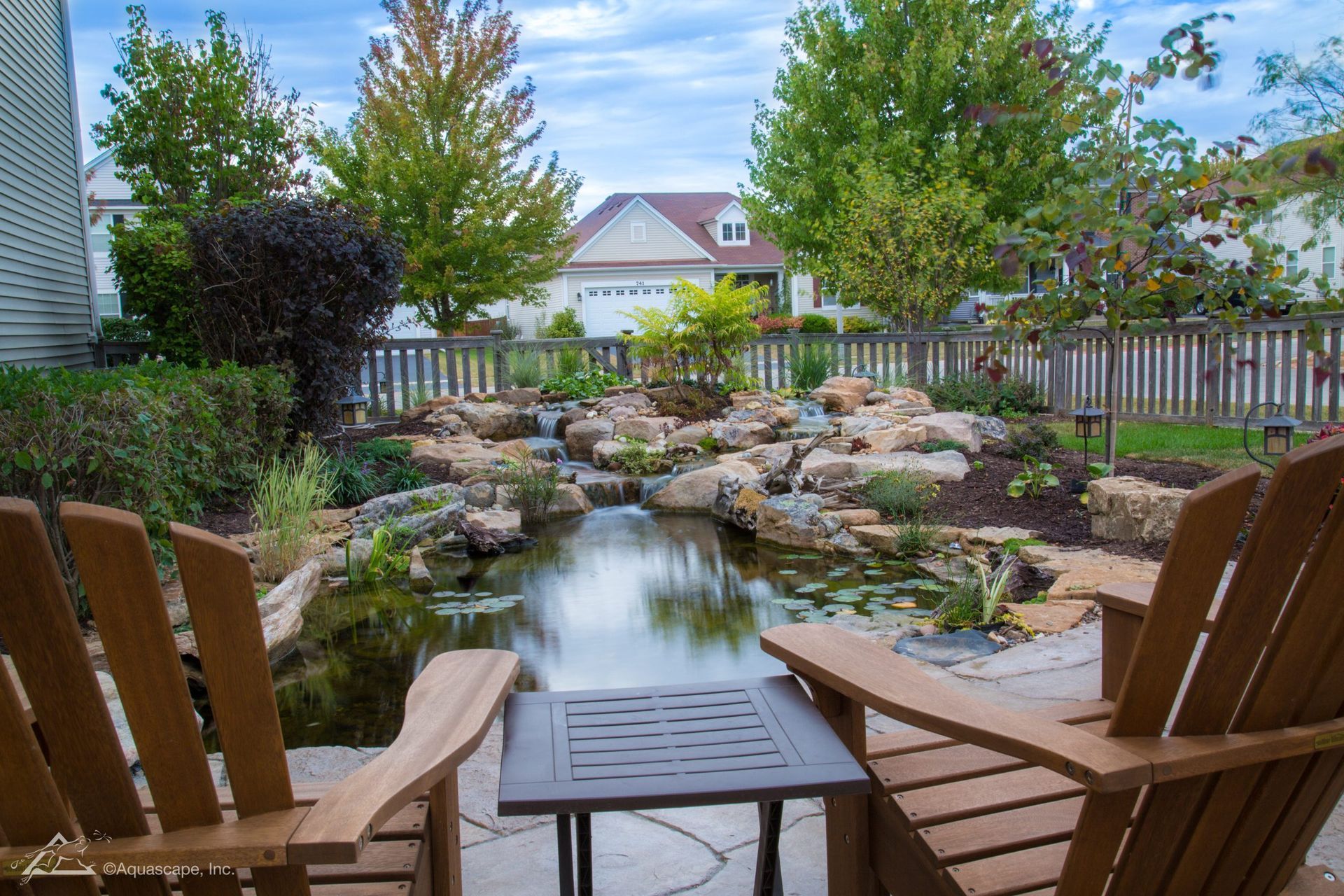 Two wooden chairs face a pond and rock garden in a backyard, with a house in the background.