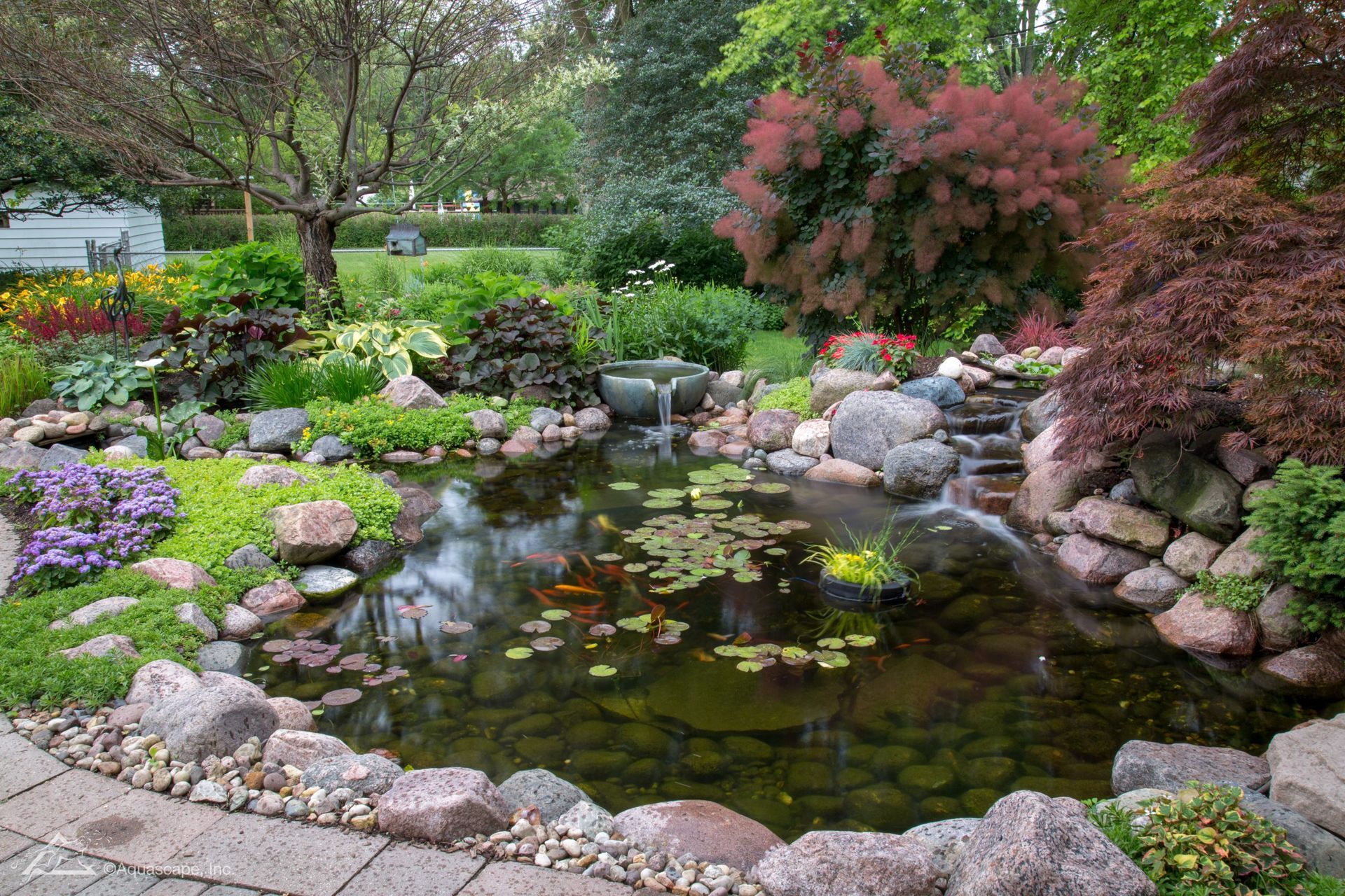 A lush garden with a pond and waterfall, surrounded by flowers, rocks, and trees.