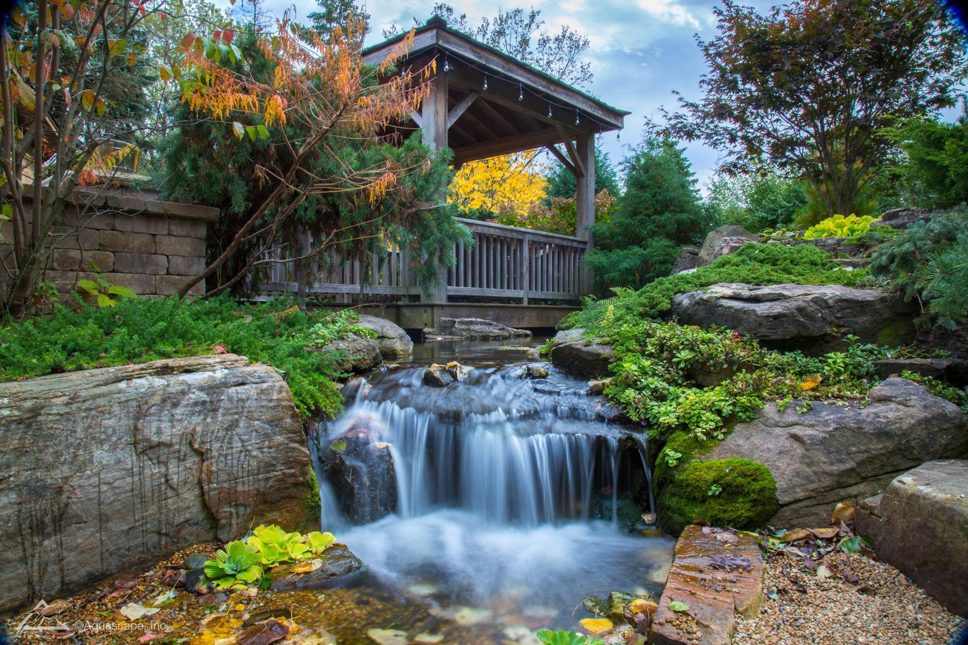 Waterfall flowing over rocks in a garden; wooden gazebo in the background, surrounded by lush plants and colorful foliage.